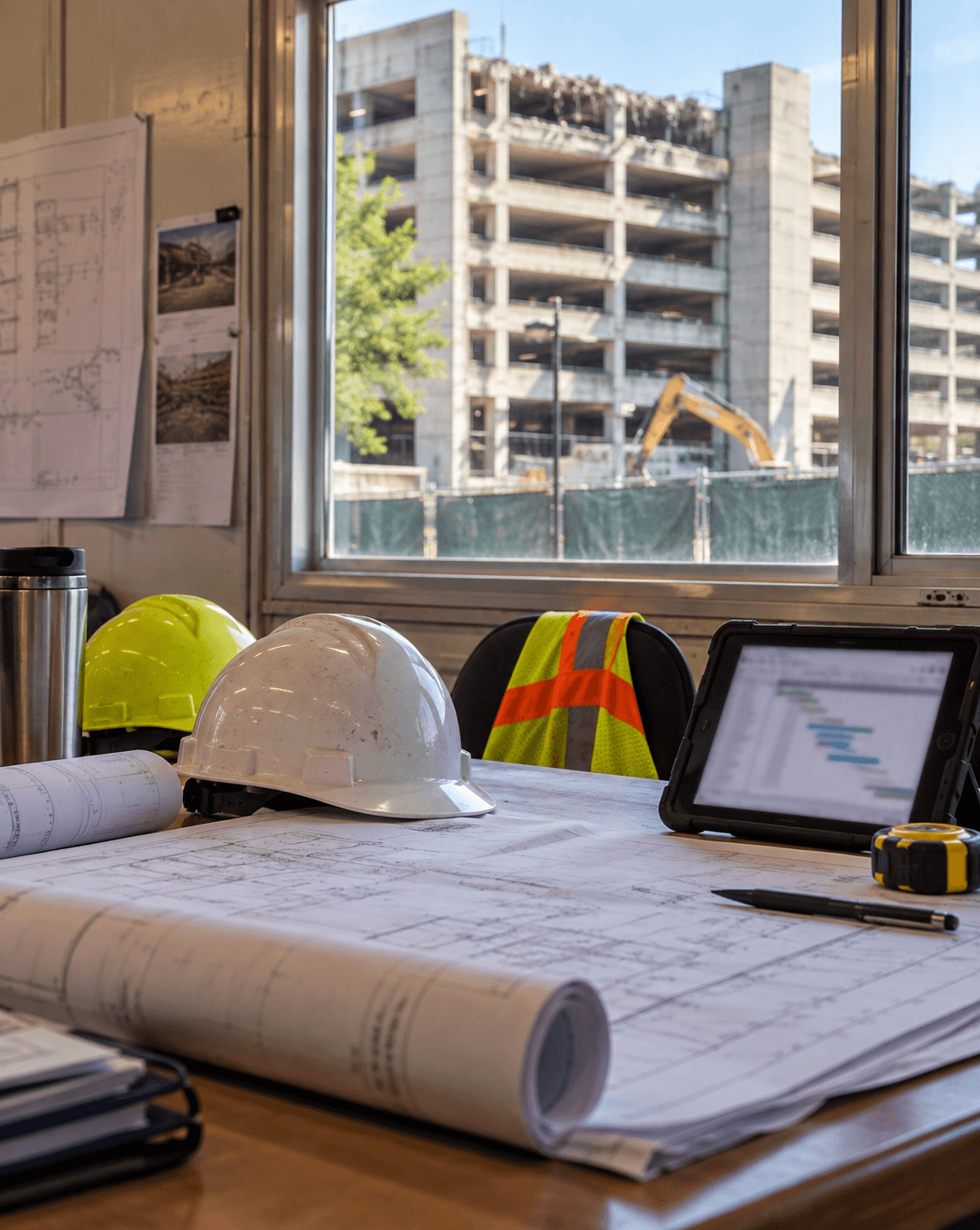 Commercial jobsite trailer table with rolled drawings, hard hats, and tablet beside a window view of a multi-level parking garage — permits and GC coordination